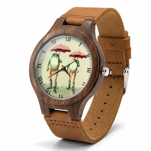 May include: A wristwatch with a dark brown wooden case and a tan leather strap. The watch face features a cream background with Roman numerals and an illustration of two frogs standing upright, wearing boots, and holding small pink umbrellas.