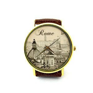 May include: A gold-tone wristwatch with a brown leather band. The watch face features a black and white photograph of the skyline of Rome, Italy. The word "Rome" is printed in cursive at the top of the watch face.