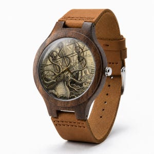 May include: A wristwatch with a dark brown wooden case and a tan leather strap. The watch face features a detailed illustration of a giant octopus attacking a sailing ship on a beige background, with thin gold hands and a silver crown on the side.