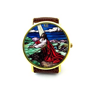 May include: A gold-tone wristwatch with a brown leather band features a stained glass image of Jesus praying in a landscape setting. The watch face has Roman numeral hour markers.