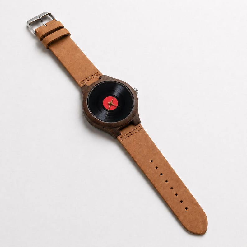 May include: A wristwatch with a brown leather band and a wooden watch face. The watch face is a black vinyl record with a red center. The watch has a silver buckle.