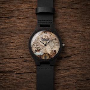May include: A wristwatch with a black wooden case and a black leather strap. The watch face features a vintage-style world map background with illustrations of a sailing ship, a treasure chest, and a compass, marked by black Roman numerals.