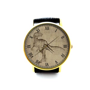 Puede incluir: Un reloj de pulsera dorado con una correa de cuero negro. La esfera del reloj presenta una ilustraci&oacute;n de estilo vintage de un dinosaurio con marcadores de hora en n&uacute;meros romanos.