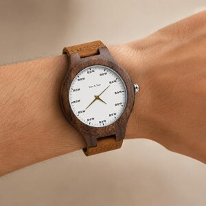 K&ouml;nnte beinhalten: Eine Holzarmbanduhr mit braunem Lederarmband. Das wei&szlig;e Zifferblatt zeigt das Wort "now" anstelle von Zahlen. Goldene Zeiger und der Text "Time Is Now" sind sichtbar.