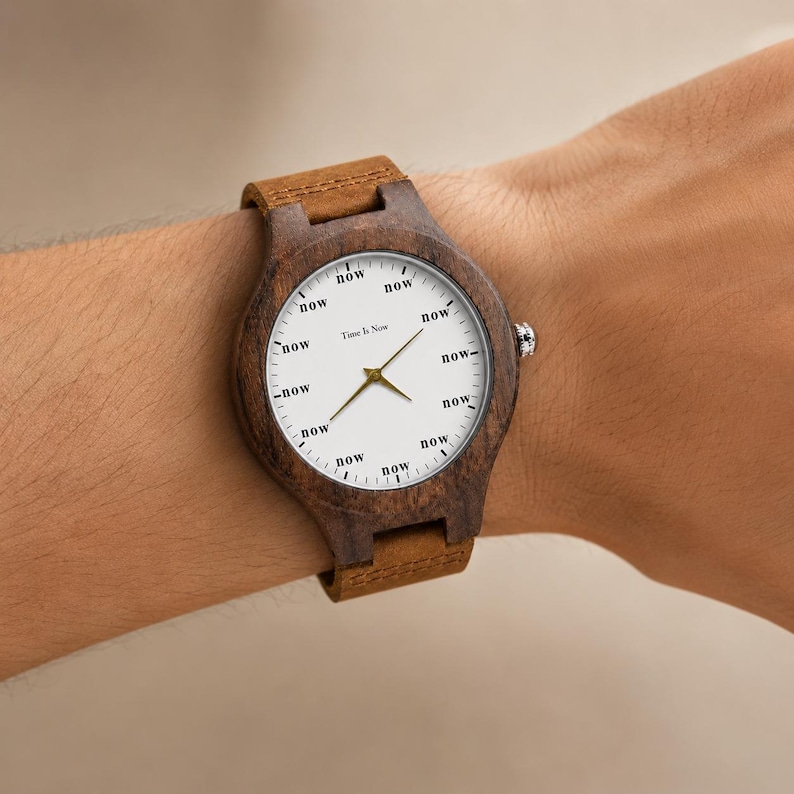 K&ouml;nnte beinhalten: Eine Holzarmbanduhr mit braunem Lederarmband. Das wei&szlig;e Zifferblatt zeigt das Wort "now" anstelle von Zahlen. Goldene Zeiger und der Text "Time Is Now" sind sichtbar.