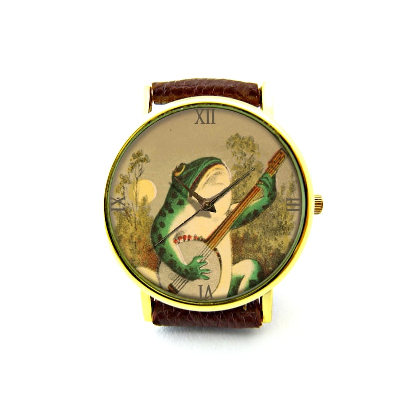 K&ouml;nnte beinhalten: Eine goldfarbene Armbanduhr mit braunem Lederarmband zeigt eine Vintage-Illustration eines gr&uuml;nen Frosches, der in einer Waldlandschaft Banjo spielt. Das Zifferblatt der Uhr hat r&ouml;mische Ziffern f&uuml;r die Stunden.