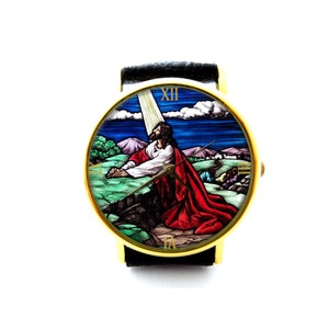 May include: A gold-tone wristwatch with a black leather band. The watch face features a stained glass image of Jesus praying in a landscape setting. The Roman numeral XII is visible on the watch face.