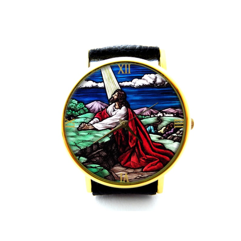 May include: A gold-tone wristwatch with a black leather band. The watch face features a stained glass image of Jesus praying in a landscape setting. The Roman numeral XII is visible on the watch face.