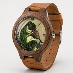 May include: A wristwatch with a dark brown wooden case and a tan leather strap. The watch face features a vintage-style illustration of a green frog playing a violin, with Roman numerals at the twelve, three, and nine o'clock positions.