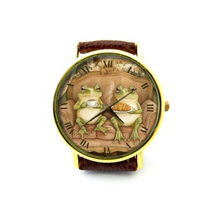 May include: A gold-tone wristwatch with a brown leather band features a whimsical illustration of two frogs sitting on a couch, one holding a cup of tea and the other holding a pastry. The watch face has Roman numeral hour markers.