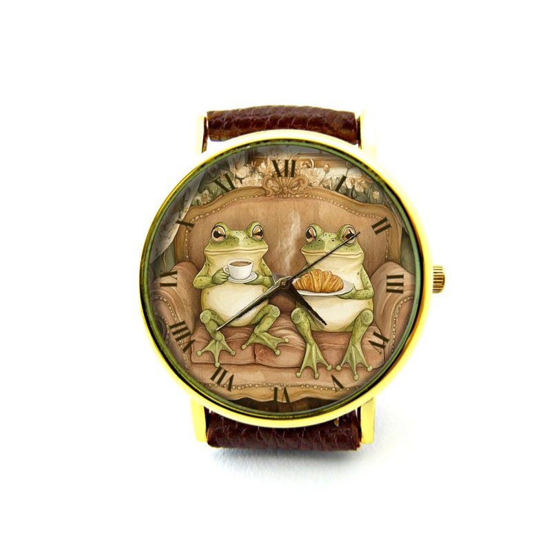 May include: A gold-tone wristwatch with a brown leather band features a whimsical illustration of two frogs sitting on a couch, one holding a cup of tea and the other holding a pastry. The watch face has Roman numeral hour markers.