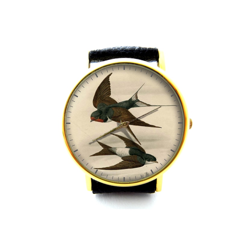 K&ouml;nnte beinhalten: Eine goldfarbene Armbanduhr mit schwarzem Lederarmband hat ein wei&szlig;es Zifferblatt mit einer Vintage-Illustration von zwei Schwalben im Flug.