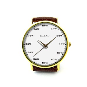 Peut inclure: Une montre-bracelet dor&eacute;e avec un cadran blanc et un bracelet en cuir marron. Le cadran de la montre affiche le mot "now" &agrave; la place des chiffres et le texte "Time Is Now". Les aiguilles sont de couleur bronze.