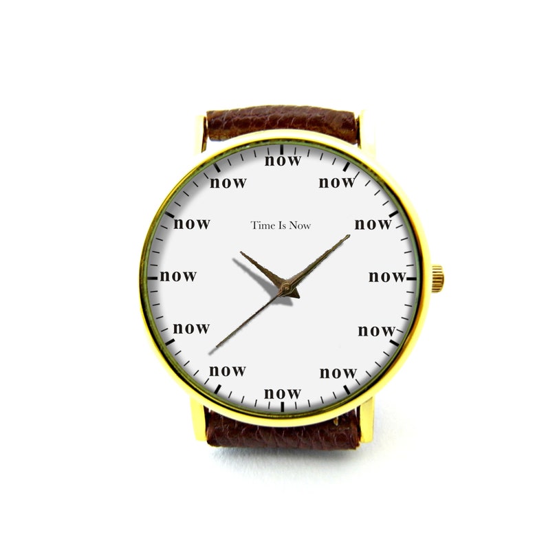 Peut inclure: Une montre-bracelet dor&eacute;e avec un cadran blanc et un bracelet en cuir marron. Le cadran de la montre affiche le mot "now" &agrave; la place des chiffres et le texte "Time Is Now". Les aiguilles sont de couleur bronze.
