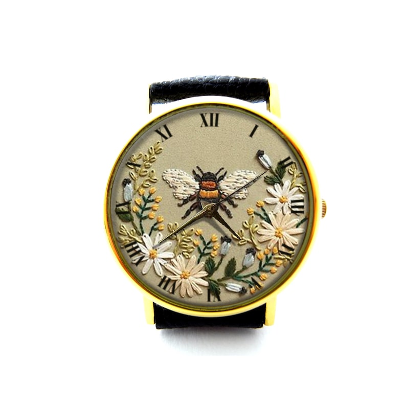 K&ouml;nnte beinhalten: Eine goldfarbene Armbanduhr mit einem schwarzen Lederarmband. Das Zifferblatt der Uhr hat einen beigefarbenen Hintergrund mit gestickten wei&szlig;en Blumen, gr&uuml;nen Bl&auml;ttern und einer Biene in der Mitte. Die r&ouml;mischen Ziffern f&uuml;r die Stunden sind am Rand des Zifferblatts angebracht.