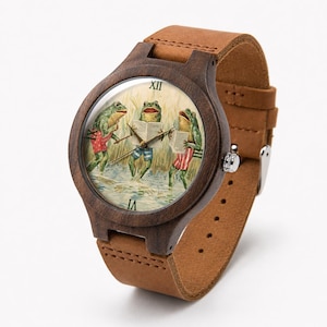 May include: A wristwatch with a dark brown wooden case and a tan leather strap. The watch face features an illustration of three frogs standing in water, each wearing patterned shorts and holding a book. Roman numerals mark the hours.