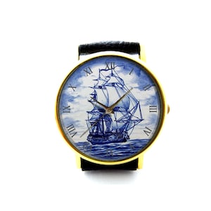 Op de afbeelding: Een gouden polshorloge met een zwart leren bandje. De wijzerplaat van het horloge heeft een blauw en wit Delfts blauw ontwerp van een zeilschip met Romeinse cijfers voor de uren.