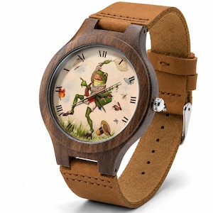 May include: A wristwatch with a dark brown wooden case and a tan leather strap. The watch face features a cream background with Roman numerals and an illustration of a green frog wearing a vest and hat, holding a net while chasing insects.