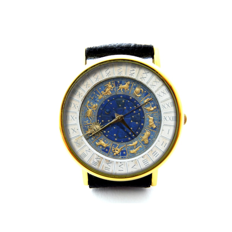 Puede incluir: Un reloj de pulsera dorado con una correa de cuero negro. La esfera del reloj tiene un fondo blanco con n&uacute;meros romanos y un centro azul con s&iacute;mbolos del zod&iacute;aco dorados.