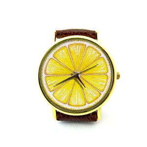 Pode incluir: Um rel&oacute;gio de pulso dourado com uma pulseira de couro marrom. O mostrador do rel&oacute;gio apresenta uma ilustra&ccedil;&atilde;o de uma fatia de lim&atilde;o amarela.