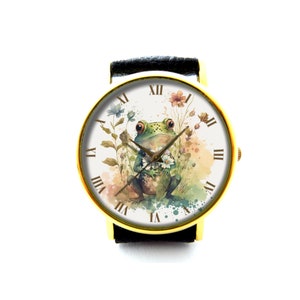 May include: A gold-tone wristwatch with a black leather band. The watch face features a watercolor illustration of a green frog holding a flower, surrounded by other flowers and greenery. The watch face also has Roman numeral hour markers.