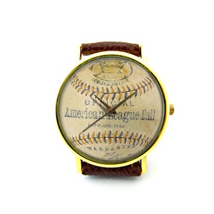 May include: A gold-tone wristwatch with a brown leather band features a vintage baseball design. The watch face shows a close-up of a baseball with the text "Official American League Ball" printed on it.