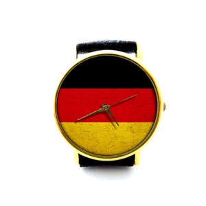 Könnte beinhalten: Eine goldene Armbanduhr mit einem schwarzen Lederarmband und einem Zifferblatt mit der deutschen Flagge. Die Zeiger der Uhr sind auf 3:00 Uhr eingestellt.