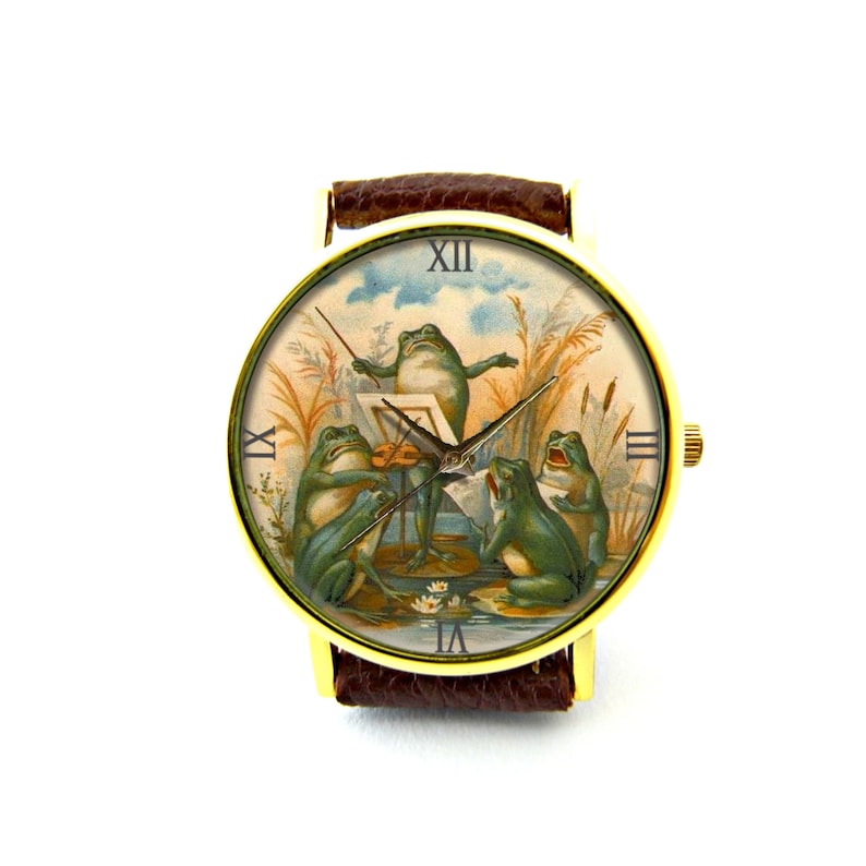 May include: A gold-tone wristwatch with a brown leather band. The watch face features a vintage illustration of four frogs playing music. The frog in the center is conducting with a baton, while the other frogs are playing a violin, a cello, and a trumpet.