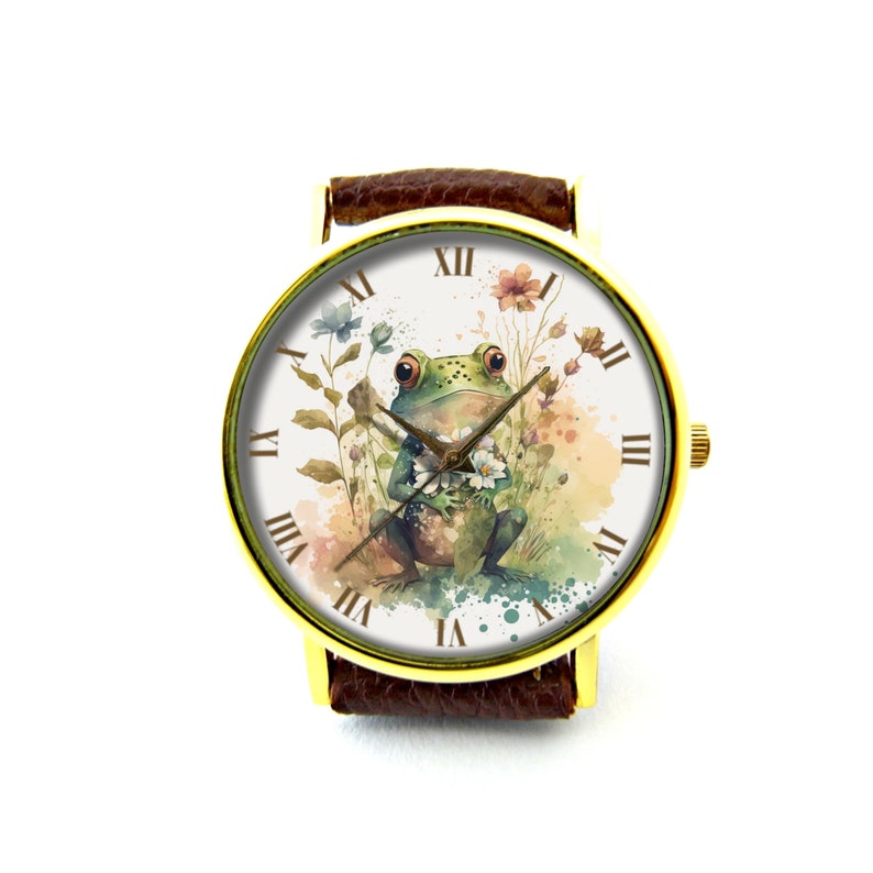 May include: A gold-tone wristwatch with a brown leather band. The watch face features a watercolor illustration of a green frog holding a flower, surrounded by other flowers and greenery. The watch face has Roman numeral hour markers.