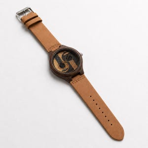 May include: A wooden wristwatch with a brown leather band. The watch face features two guitar designs, one light and one dark, in a yin-yang style. The band is light brown and has a silver buckle.