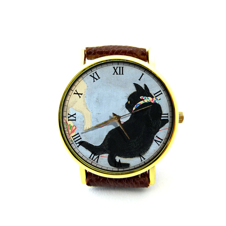 May include: A gold-tone wristwatch with a brown leather band features a black cat with a colorful ribbon playing with a red string. The watch face has Roman numeral hour markers and a blue background.