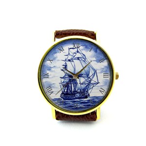 Op de afbeelding: Een gouden polshorloge met een bruin leren bandje. De wijzerplaat van het horloge heeft een blauwe en witte illustratie van een zeilschip met Romeinse cijfers voor de uren.