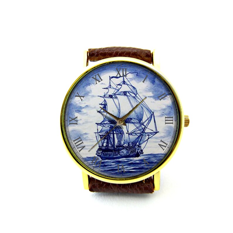Op de afbeelding: Een gouden polshorloge met een bruin leren bandje. De wijzerplaat van het horloge heeft een blauwe en witte illustratie van een zeilschip met Romeinse cijfers voor de uren.