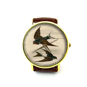 K&ouml;nnte beinhalten: Eine goldfarbene Armbanduhr mit braunem Lederarmband hat ein wei&szlig;es Zifferblatt mit einer Vintage-Illustration von zwei Schwalben im Flug.