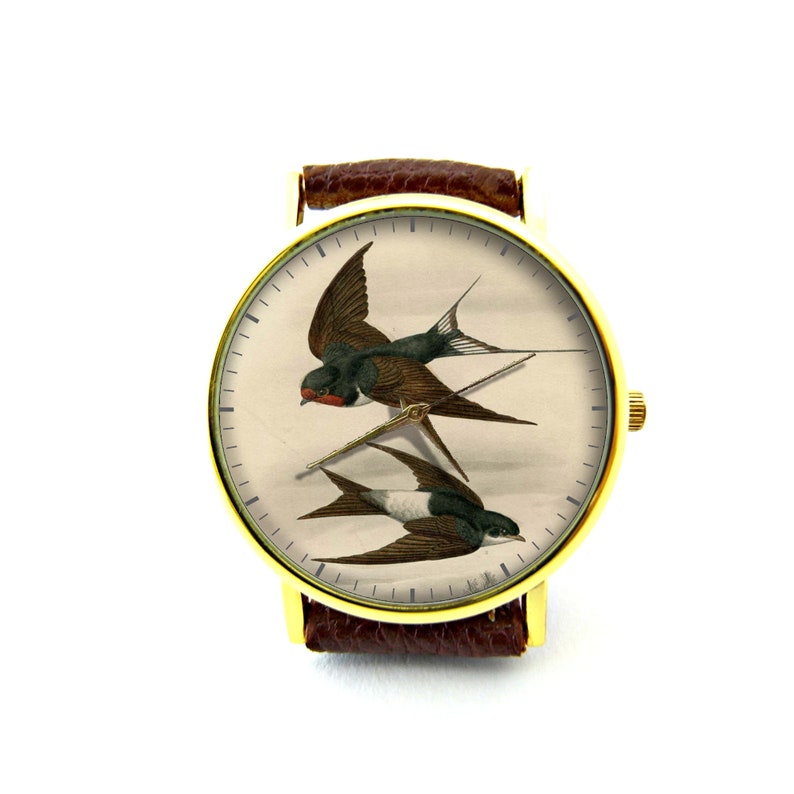 K&ouml;nnte beinhalten: Eine goldfarbene Armbanduhr mit braunem Lederarmband hat ein wei&szlig;es Zifferblatt mit einer Vintage-Illustration von zwei Schwalben im Flug.