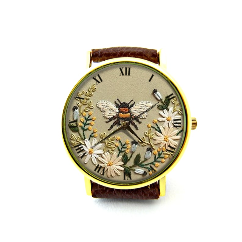 K&ouml;nnte beinhalten: Eine goldfarbene Armbanduhr mit einem braunen Lederarmband. Das Zifferblatt der Uhr zeigt ein gesticktes Design einer Biene, die von Blumen umgeben ist.