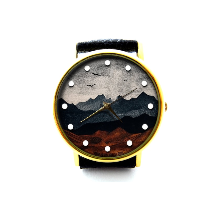 May include: A gold-tone wristwatch with a black leather band and a white face. The face features a graphic of a mountain range with a brown and blue color scheme. The watch has white hour markers and a black hour and minute hand.
