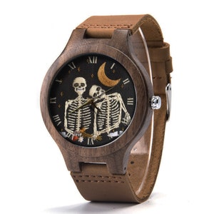 May include: A brown wooden wristwatch with a brown leather band. The watch face features an embroidered design of two skeletons holding each other, with a crescent moon and stars in the background.