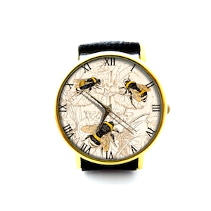 May include: A gold-tone wristwatch with a black leather strap. The watch face features a white background with a floral design and three bees. Roman numeral hours are displayed around the edge of the watch face.