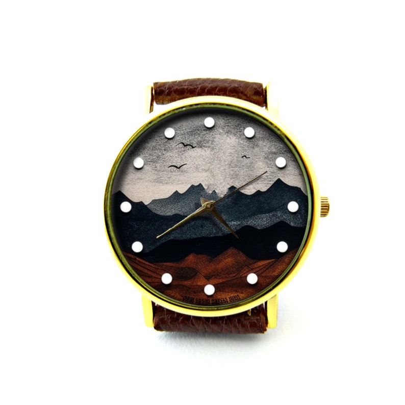 May include: A gold-tone wristwatch with a brown leather band. The watch face features a landscape design with mountains, sky, and birds. The watch face has white dots for hour markers.