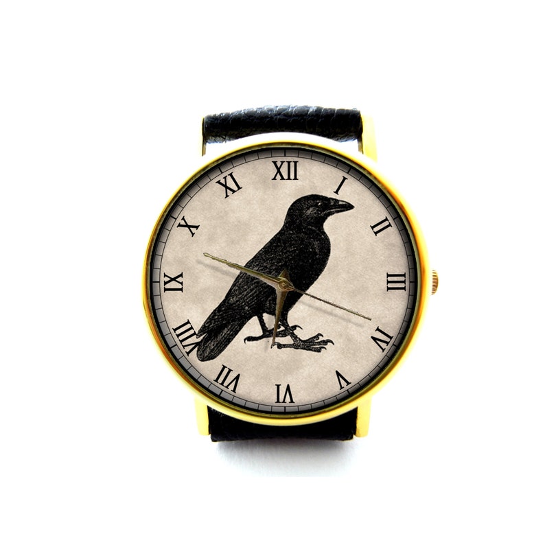 May include: A gold-tone wristwatch with a black leather band and a white face featuring a black raven illustration and Roman numeral hour markers.