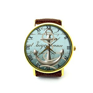 K&ouml;nnte beinhalten: Eine goldfarbene Armbanduhr mit einem braunen Lederarmband. Das Zifferblatt der Uhr hat einen blauen und wei&szlig;en Hintergrund mit einem silbernen Anker- und Seildesign. Das Zifferblatt der Uhr hat auch r&ouml;mische Ziffern als Stundenmarkierungen und den Text "Carl Zeigmann".