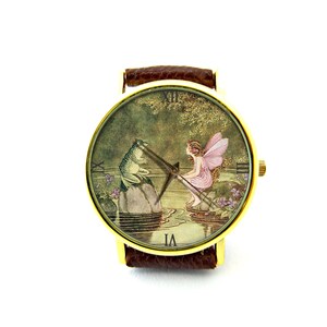 May include: A gold-tone wristwatch with a brown leather band. The watch face features a vintage illustration of a fairy with pink wings sitting on a rock by a pond, talking to a green frog.