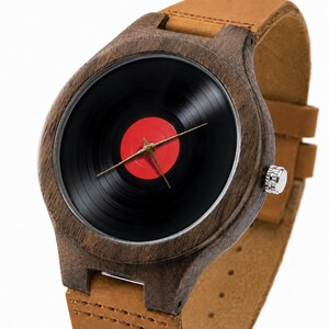 May include: A wooden wristwatch with a black vinyl record face and a red center. The watch has gold-colored hands and a brown leather band. The watch case is made of dark wood.