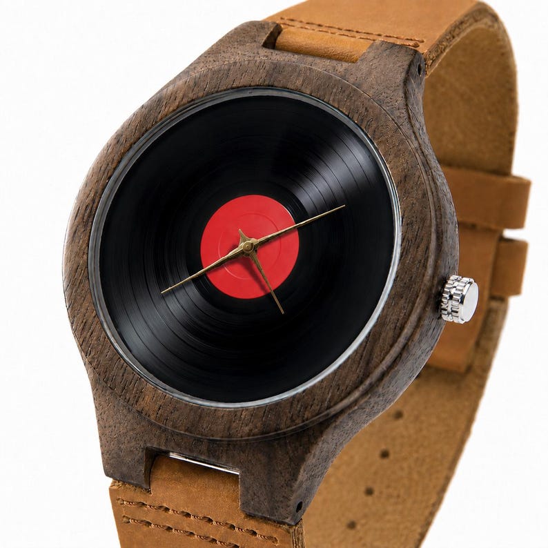 May include: A wooden wristwatch with a black vinyl record face and a red center. The watch has gold-colored hands and a brown leather band. The watch case is made of dark wood.