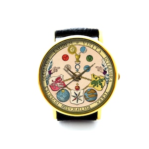 以下が含まれることがあります： 黒い革のバンドが付いたゴールドトーンの腕時計。時計の文字盤には、錬金術のシンボルと「RECTIFICANDO INVENTUS OCCULTUM LAPIDEM VISITA INTERIORA TERRAE」というテキストが描かれたヴィンテージのイラストが描かれています。