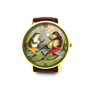 May include: A gold-tone wristwatch with a brown leather band features a vintage illustration of two frogs boxing. The watch face has Roman numeral hour markers.