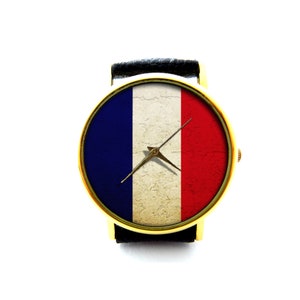 Puede incluir: Un reloj de pulsera dorado con una correa de cuero negro y una esfera con la bandera de Francia. La esfera del reloj tiene un fondo blanco con una franja azul a la izquierda y una franja roja a la derecha.