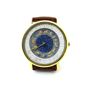Puede incluir: Un reloj de pulsera dorado con una correa de cuero marr&oacute;n. La esfera del reloj tiene un fondo blanco con n&uacute;meros romanos y un centro azul con s&iacute;mbolos del zod&iacute;aco dorados.