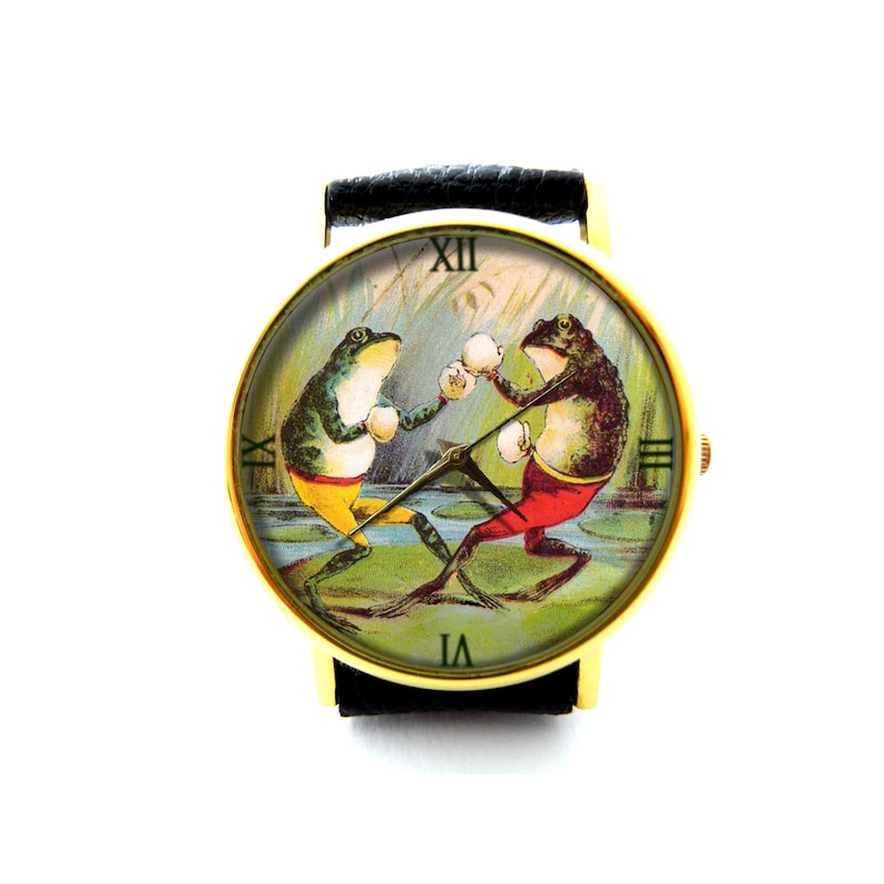 May include: A gold-tone wristwatch with a black leather band features a colorful illustration of two frogs boxing. The watch face has Roman numeral hour markers and a white background.